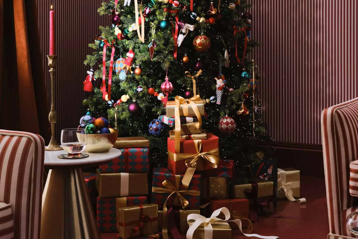 A Christmas tree hung with red ribbons and bright baubles above a pile of presents wrapped in gold and harlequin paper, between two striped armchairs