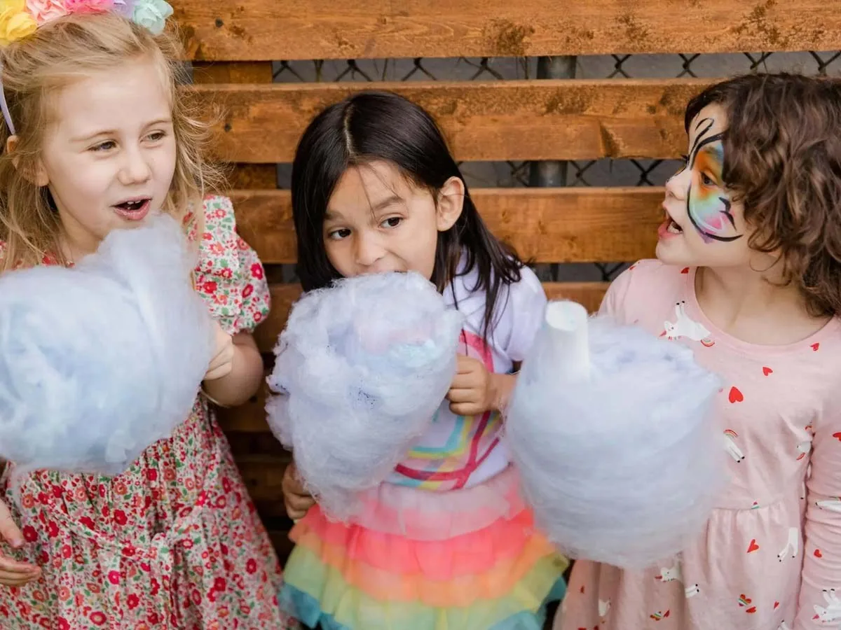 Three young children holding large sticks of candyfloss, one with face paint