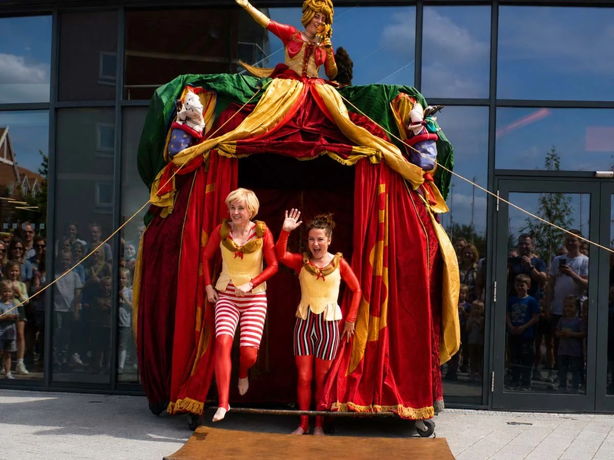Two performers in red and gold costumes stepping out of a mobile puppet theatre in front of a crowd