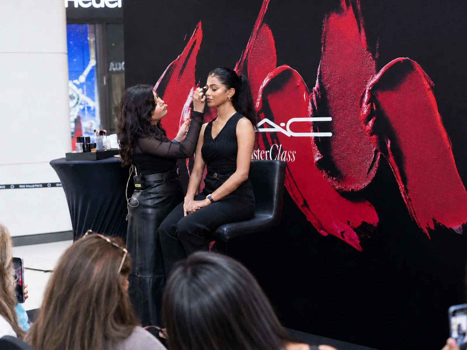 A make-up artist giving a masterclass demonstration to a seated guest in front of an audience
