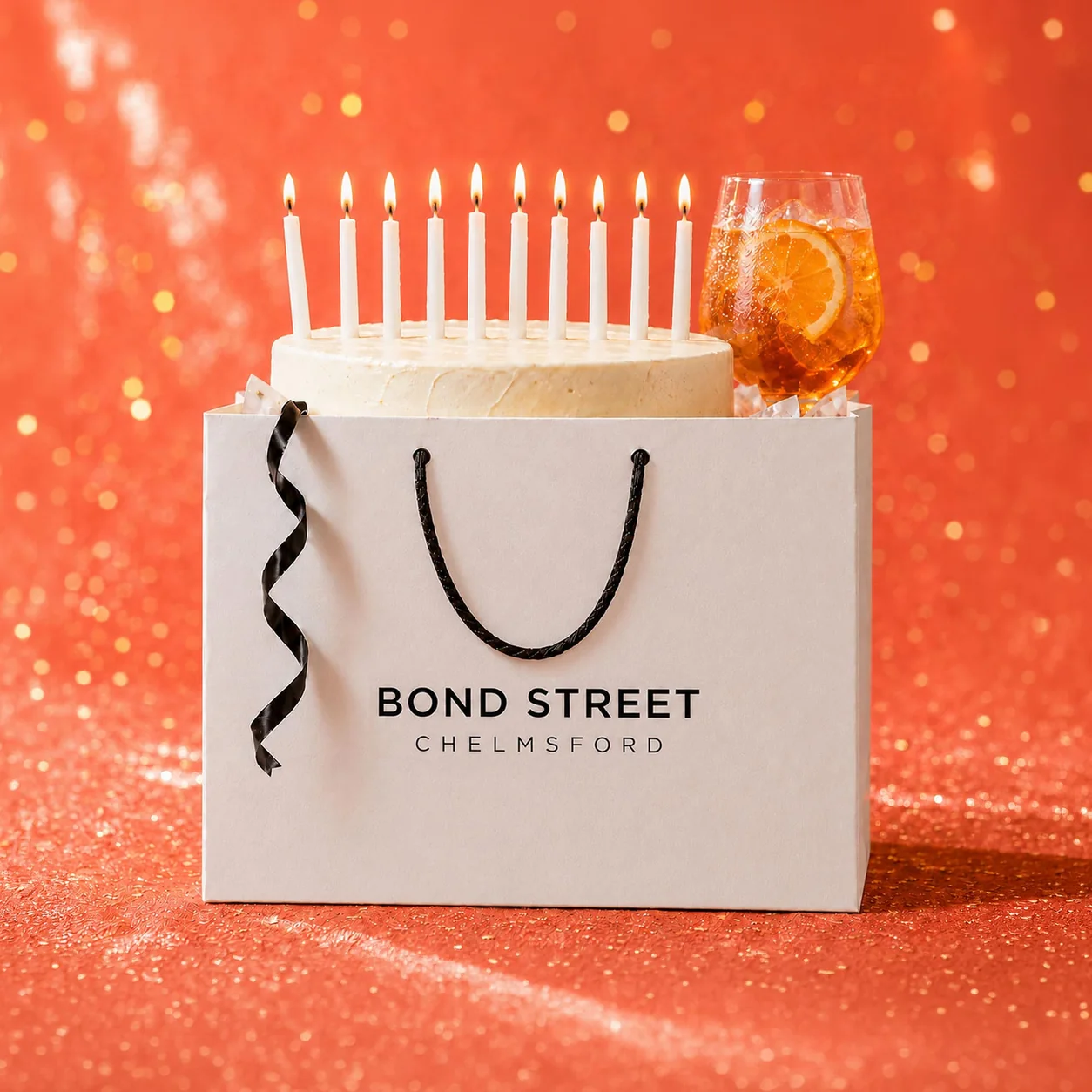 A Bond Street Chelmsford shopping bag with a birthday cake inside it, ten lit candles and an Aperol spritz, against a red background