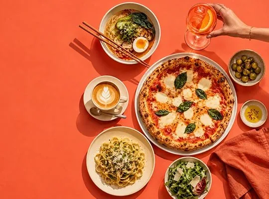 An overhead spread of pizza, pasta, ramen, coffee and olives on an orange background, with a hand holding an Aperol spritz