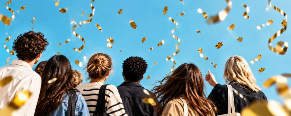 Four friends looking up as gold confetti and streamers fall against a blue sky